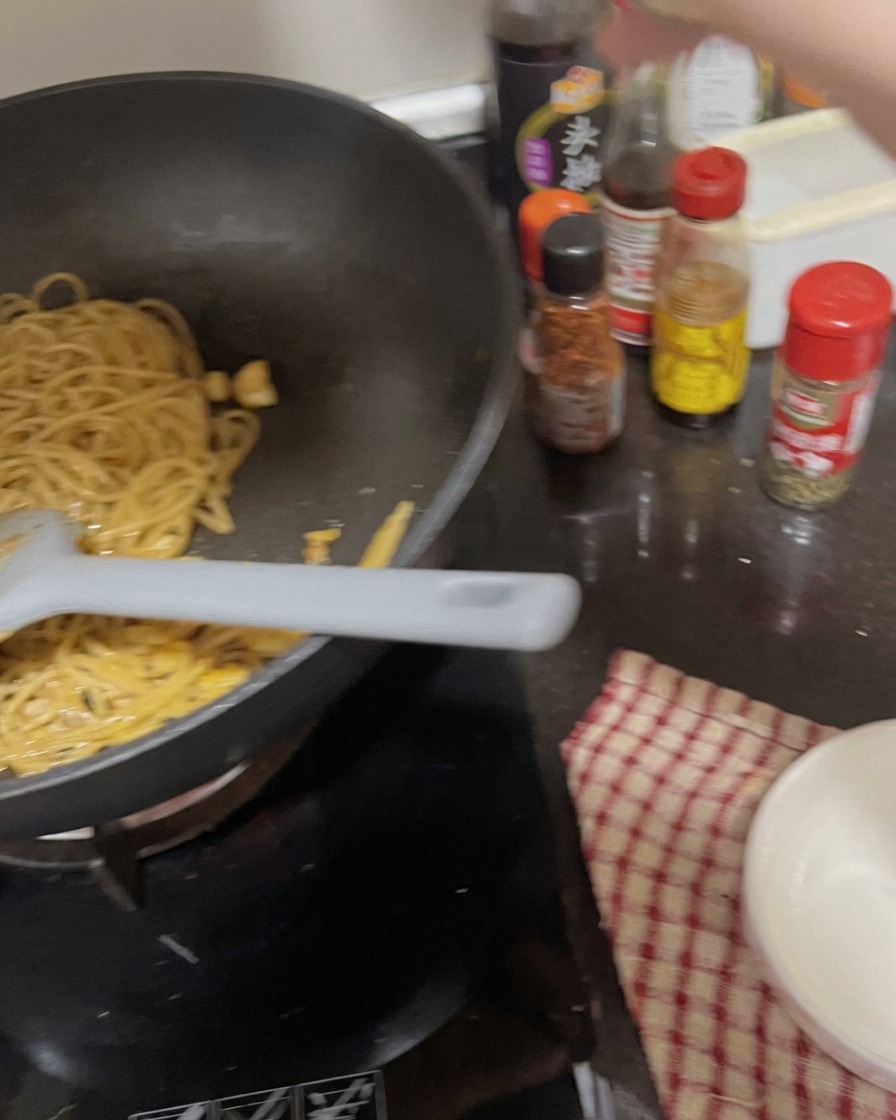 A hand shaking a seasoning container over a pan of cooked spaghetti noodles mixed with garlic.