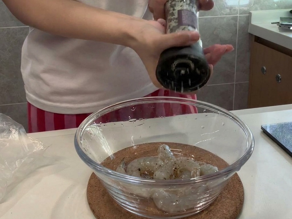 Fresh black pepper being ground over a glass bowl of raw, peeled shrimp.
