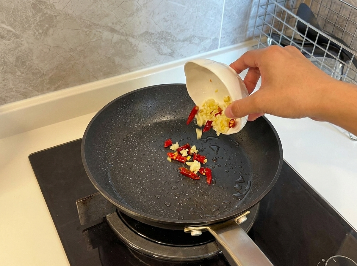 Sliced garlic and dried red chilies being poured from a small bowl into a hot, oiled pan.