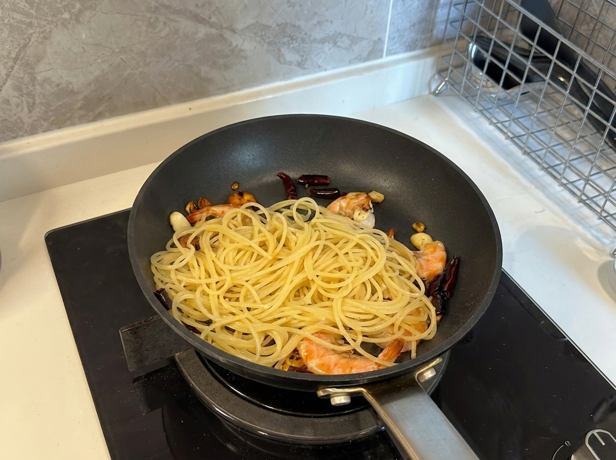 Cooked spaghetti being added to a pan filled with cooked shrimp, garlic, and dried chilies.