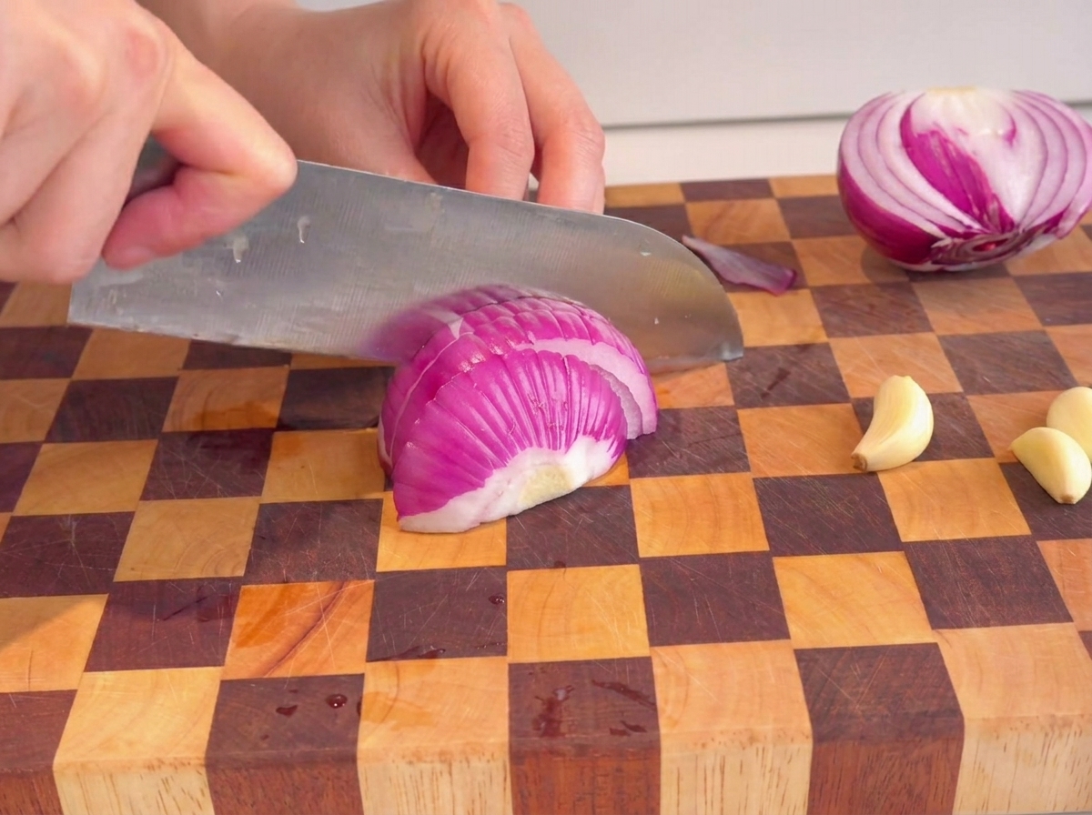 Hands using a knife to chop a red onion on a checkered cutting board.