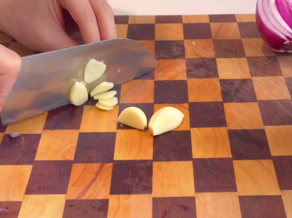Hands slicing peeled garlic cloves on a checkered wooden cutting board.