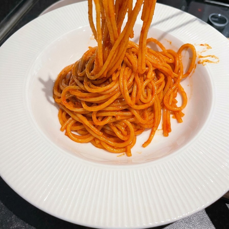Metal chopsticks lifting and twirling bright red, sauce-coated spaghetti onto a white grooved serving bowl.