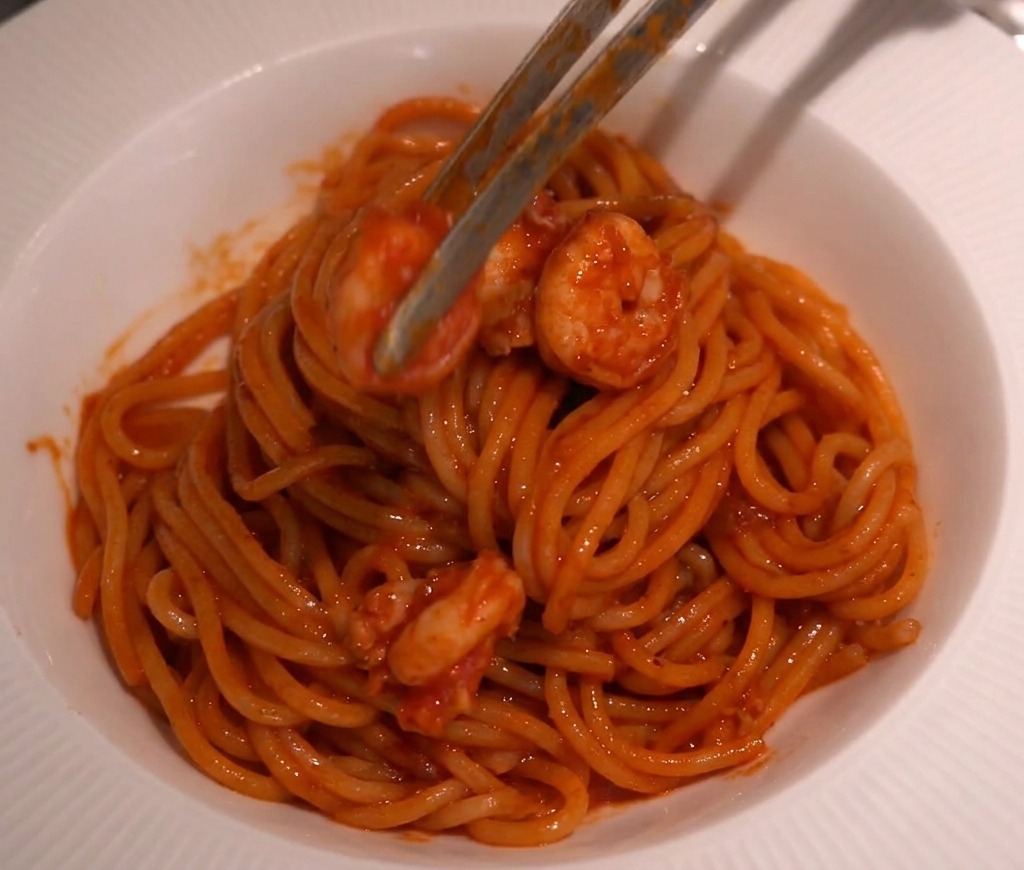 Kitchen tongs carefully placing sauced shrimp on top of a twirled mound of spaghetti in a white serving bowl.