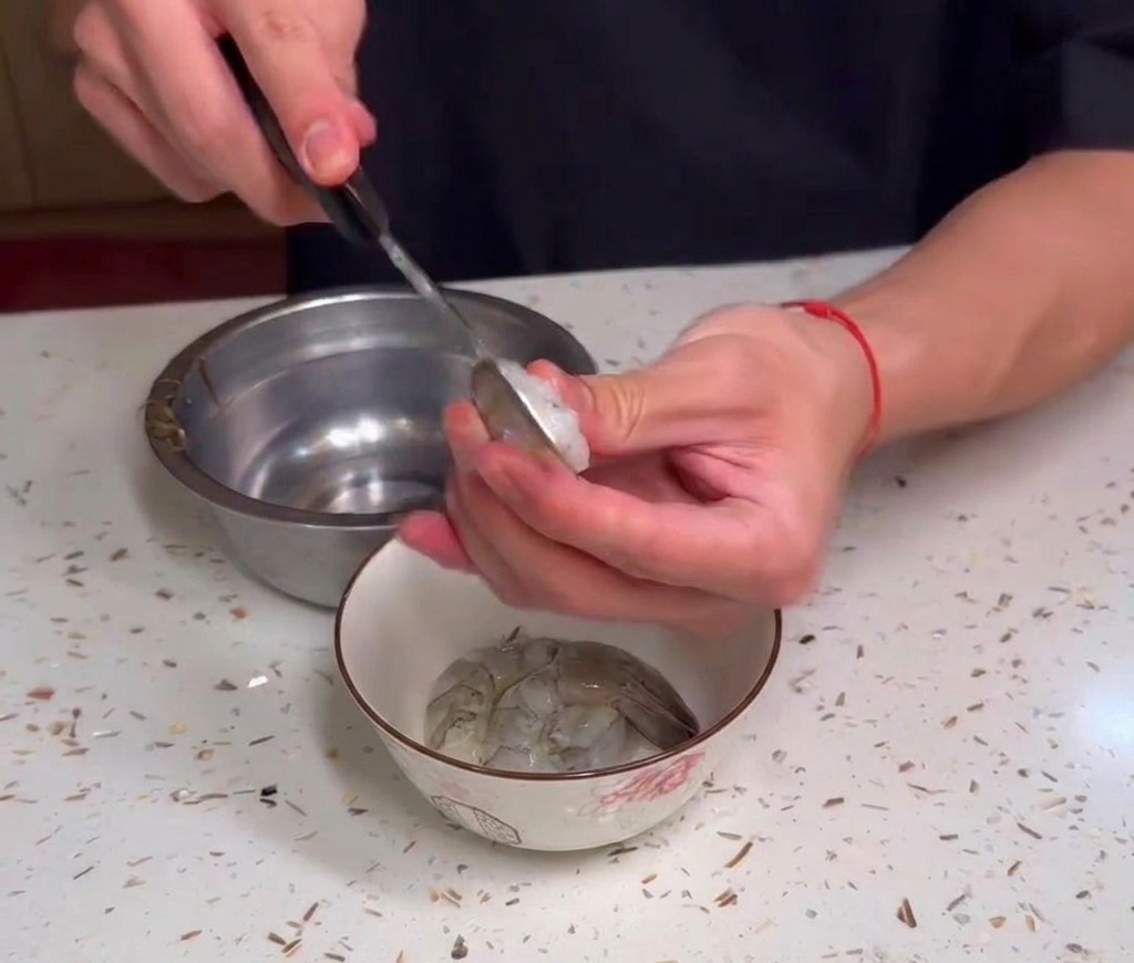 Hands holding a utensil to clean raw shrimp over a small bowl on a speckled countertop.