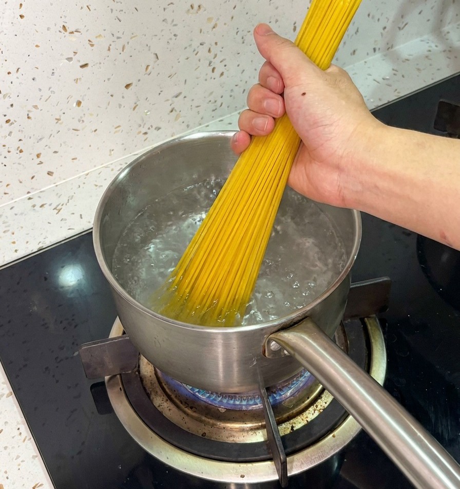 A hand firmly holding a bunch of dry spaghetti, placing it into a stainless steel pot of actively boiling water.