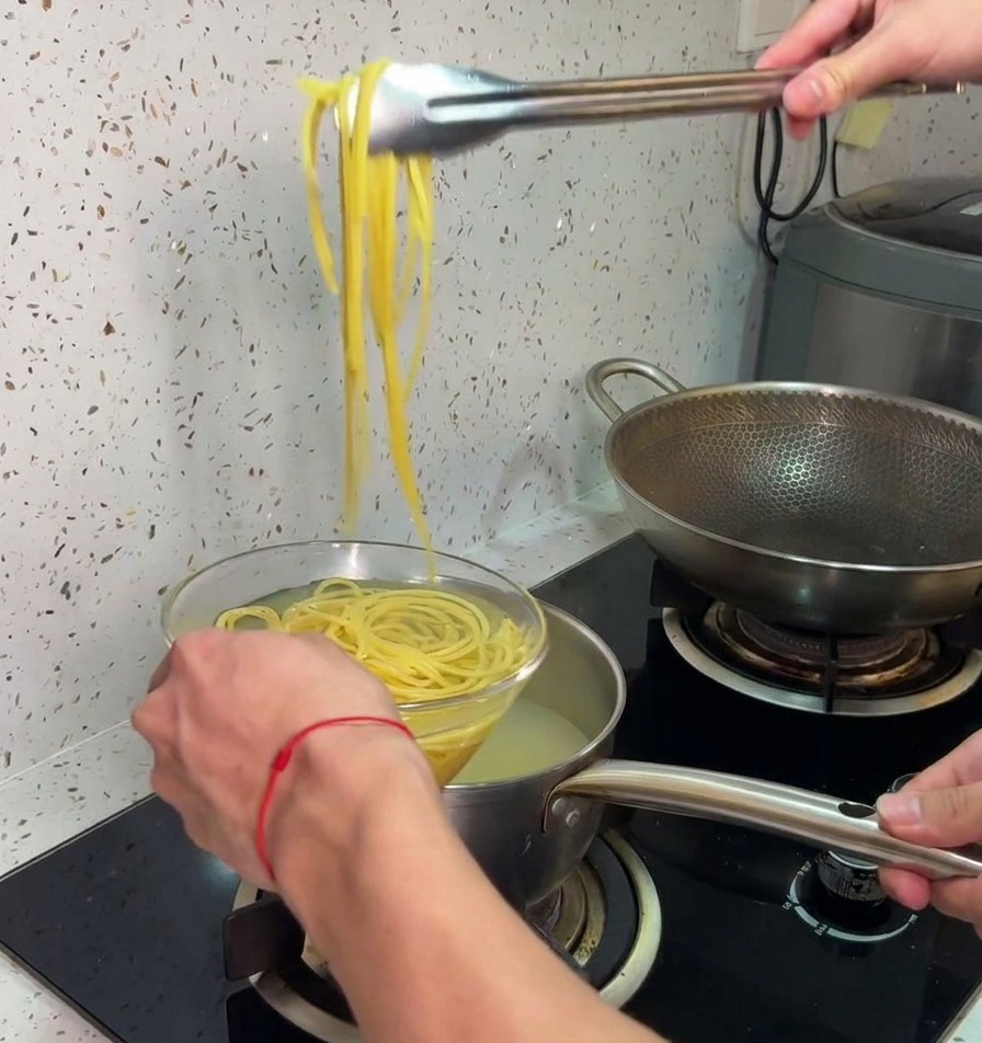 Metal tongs lifting cooked yellow spaghetti out of a hot pot and transferring it directly into a clear glass bowl of cold water.