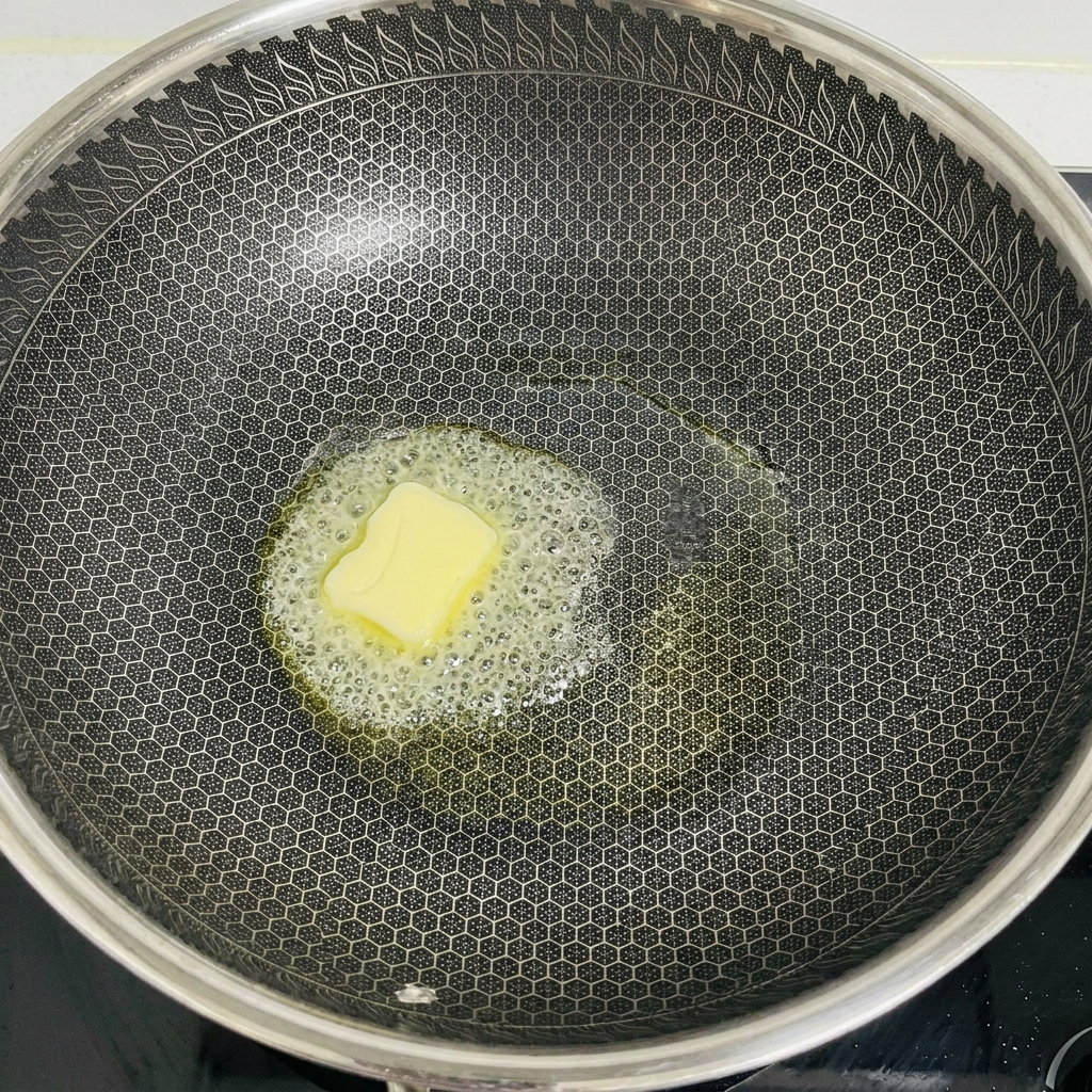 A square pat of pale yellow butter melting and bubbling in the center of a large, dark, honeycomb-patterned wok.