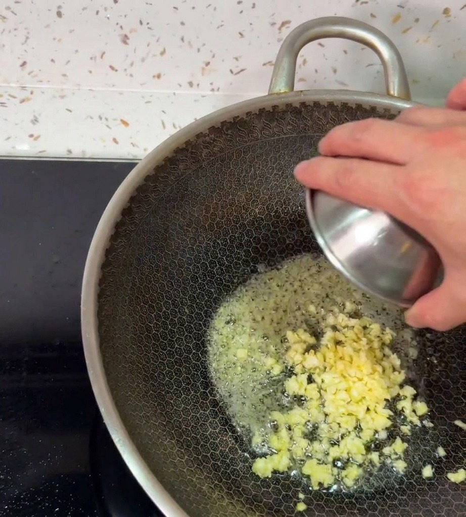 A hand tipping a small metal bowl to drop freshly minced garlic into the bubbling melted butter inside a hot wok.