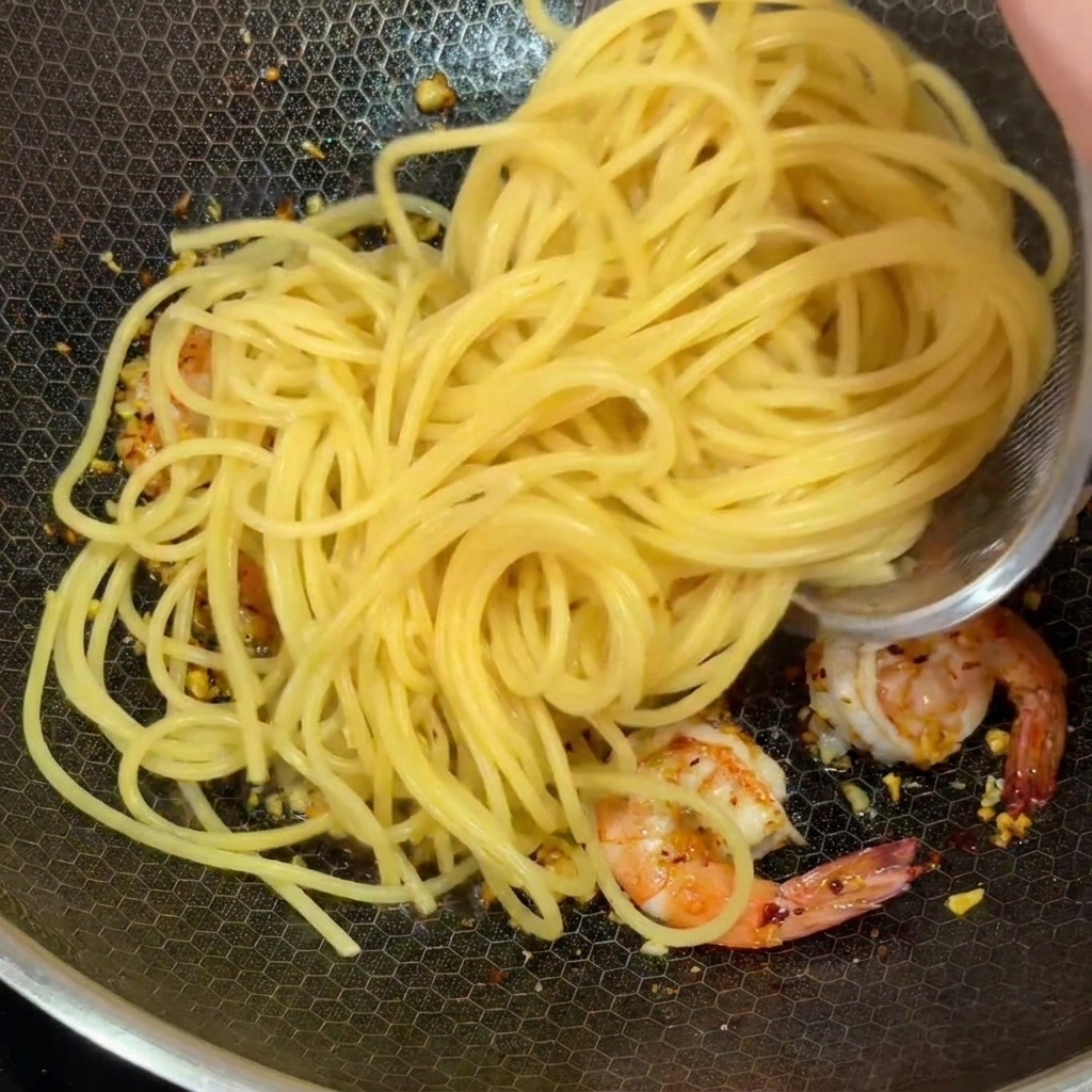Cooked spaghetti noodles being added to a wok with pink shrimp and minced garlic.