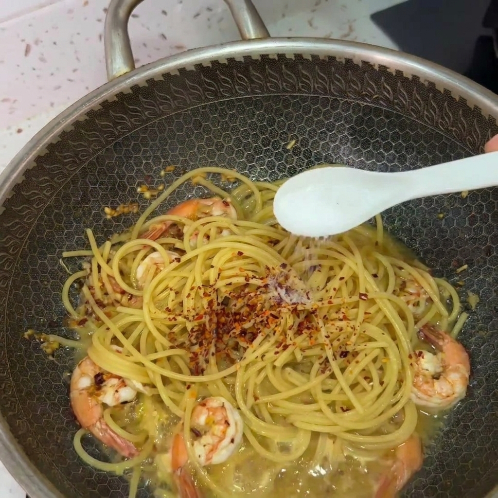 A white spoon sprinkling salt over spaghetti and shrimp in a wok, topped with red chili flakes.