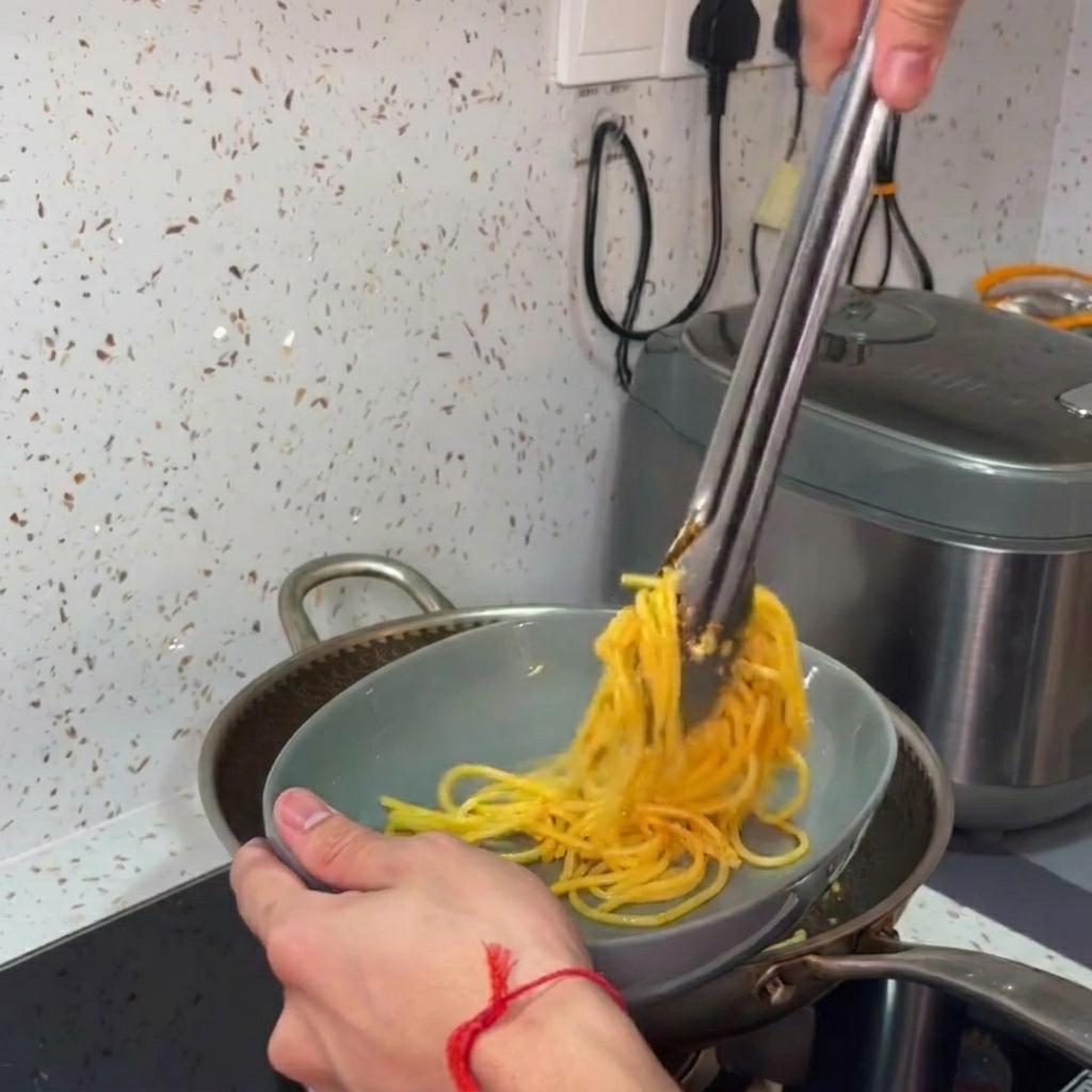 Hands using metal tongs to transfer and twist hot, seasoned spaghetti from a cooking pan into a grey serving bowl.
