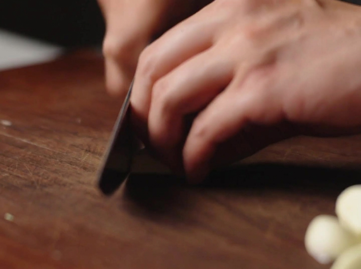 A knife slicing peeled garlic cloves on a dark wooden cutting board.