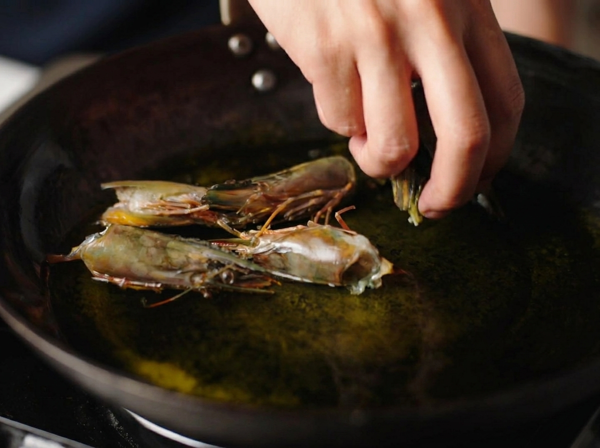 A hand placing raw shrimp heads into a wok filled with shimmering hot oil.