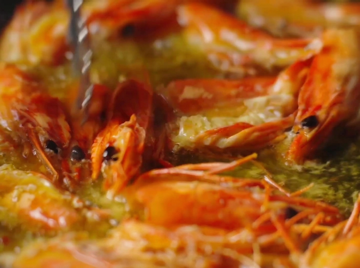 Tongs pressing down on vibrant orange shrimp heads frying in bubbling hot oil.