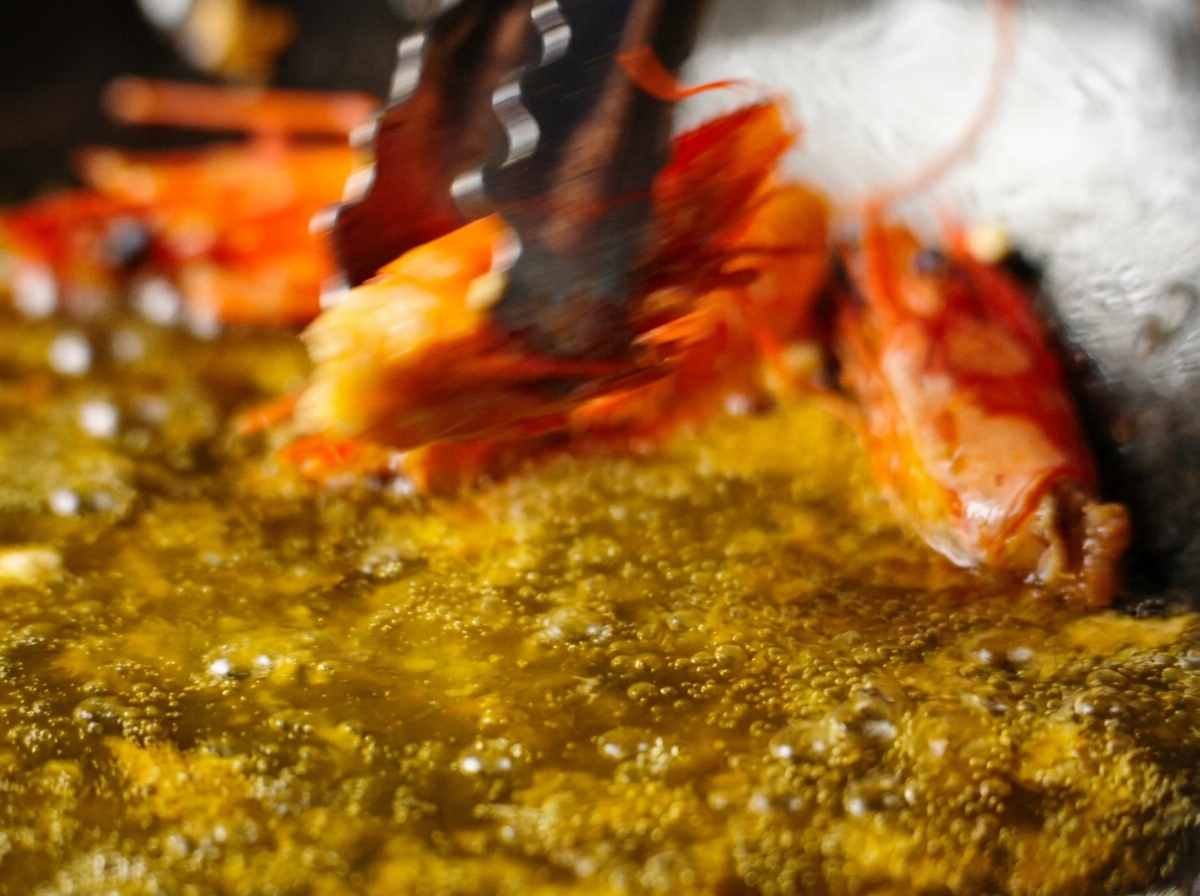 Tongs lifting a fried shrimp head out of a pan of bubbling, orange-tinted infused oil.