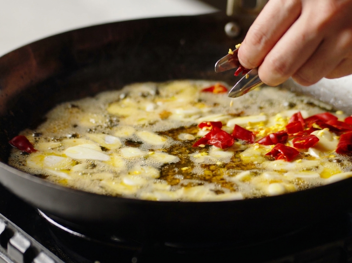 A hand dropping pieces of dried red chili into a wok with frying garlic and seasoned oil.