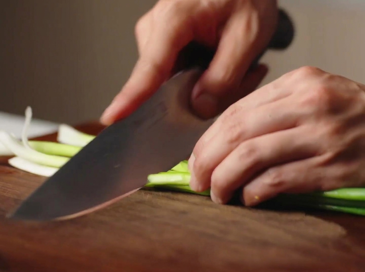 Hands using a chefs knife to chop fresh green scallions into segments on a wooden cutting board.