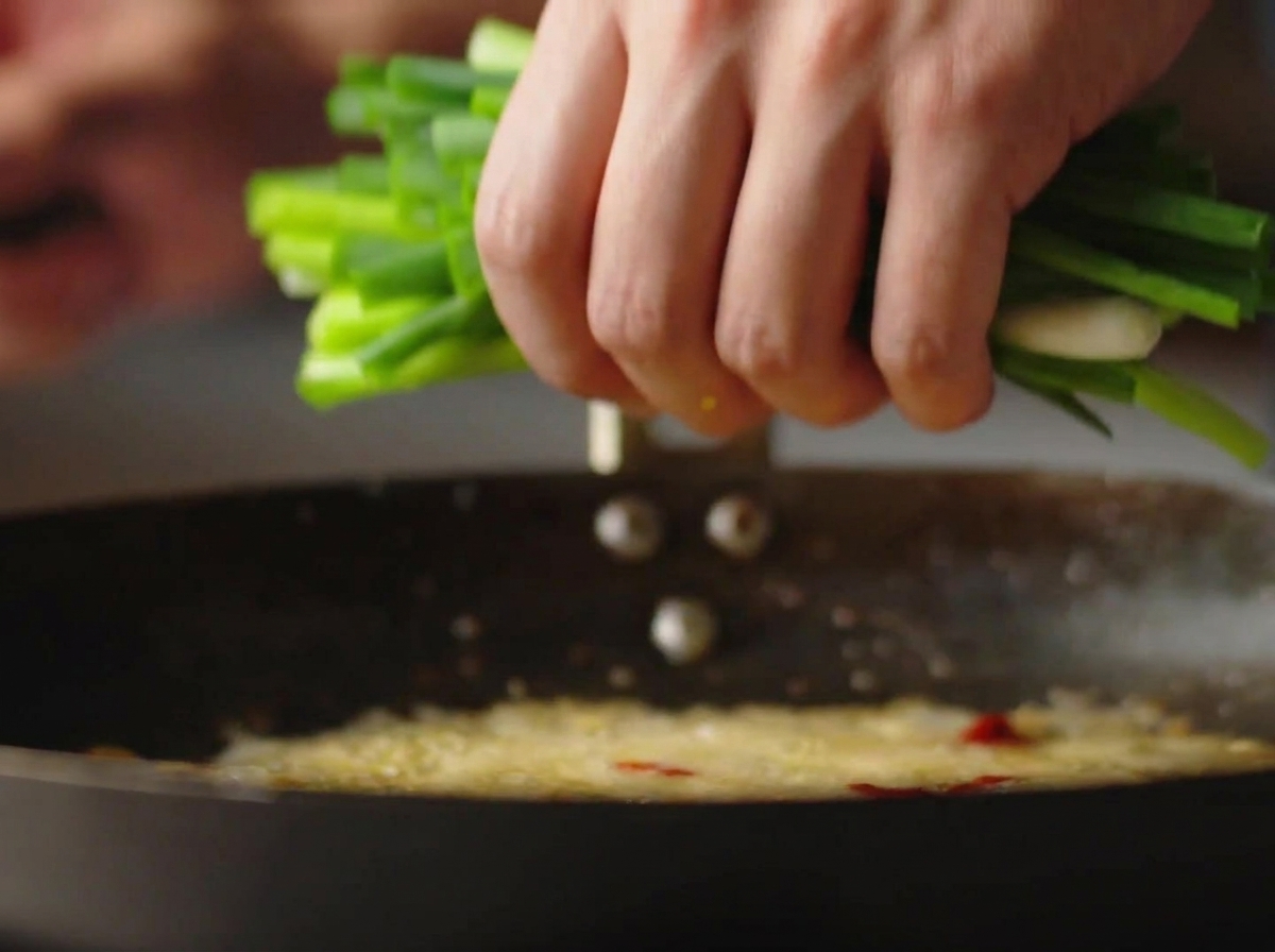 A hand dropping a large bunch of freshly chopped green scallions into a hot wok with bubbling oil.