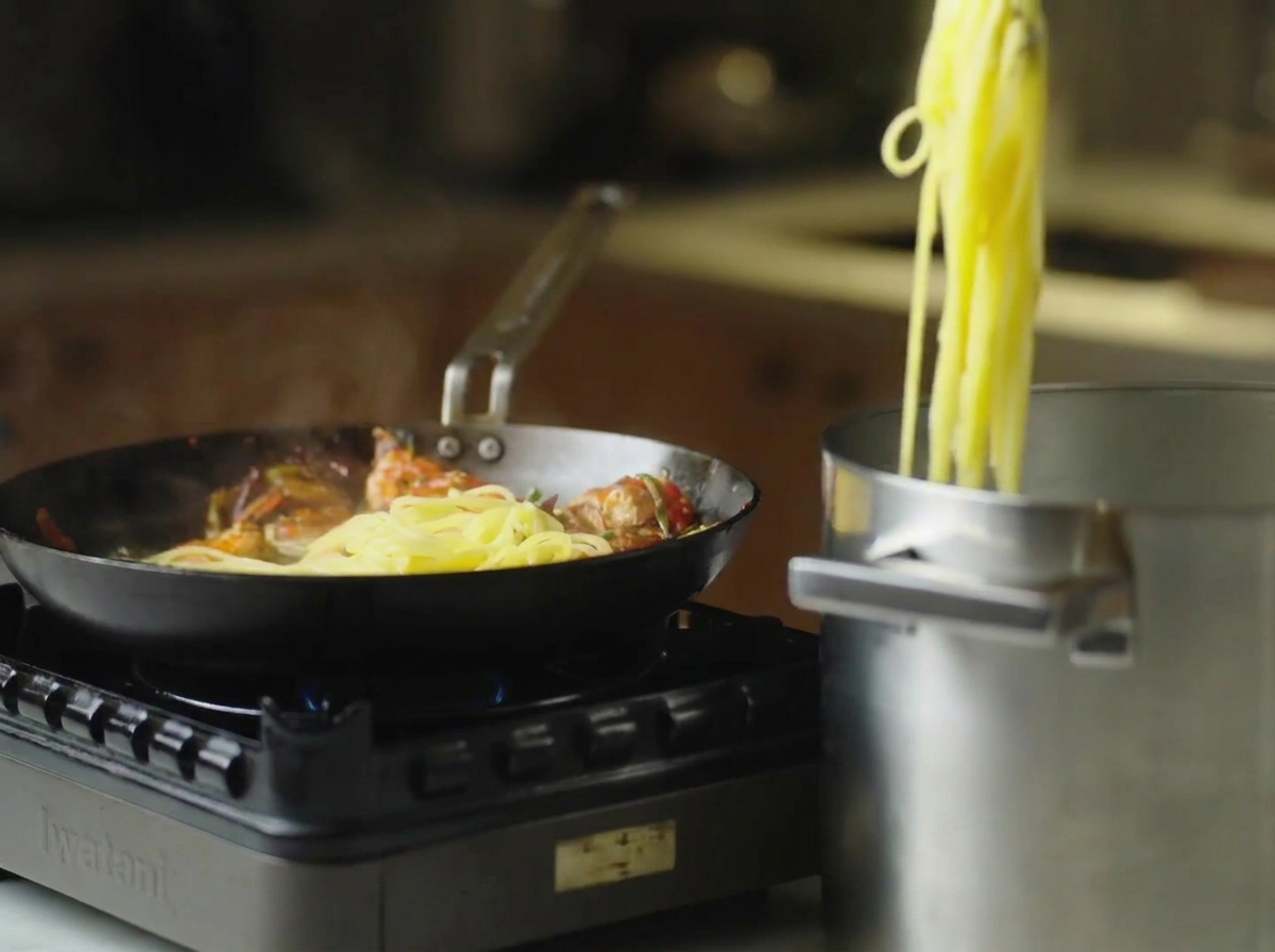 Tongs lifting hot cooked spaghetti out of a tall pot and moving it toward a skillet containing cooked shrimp.