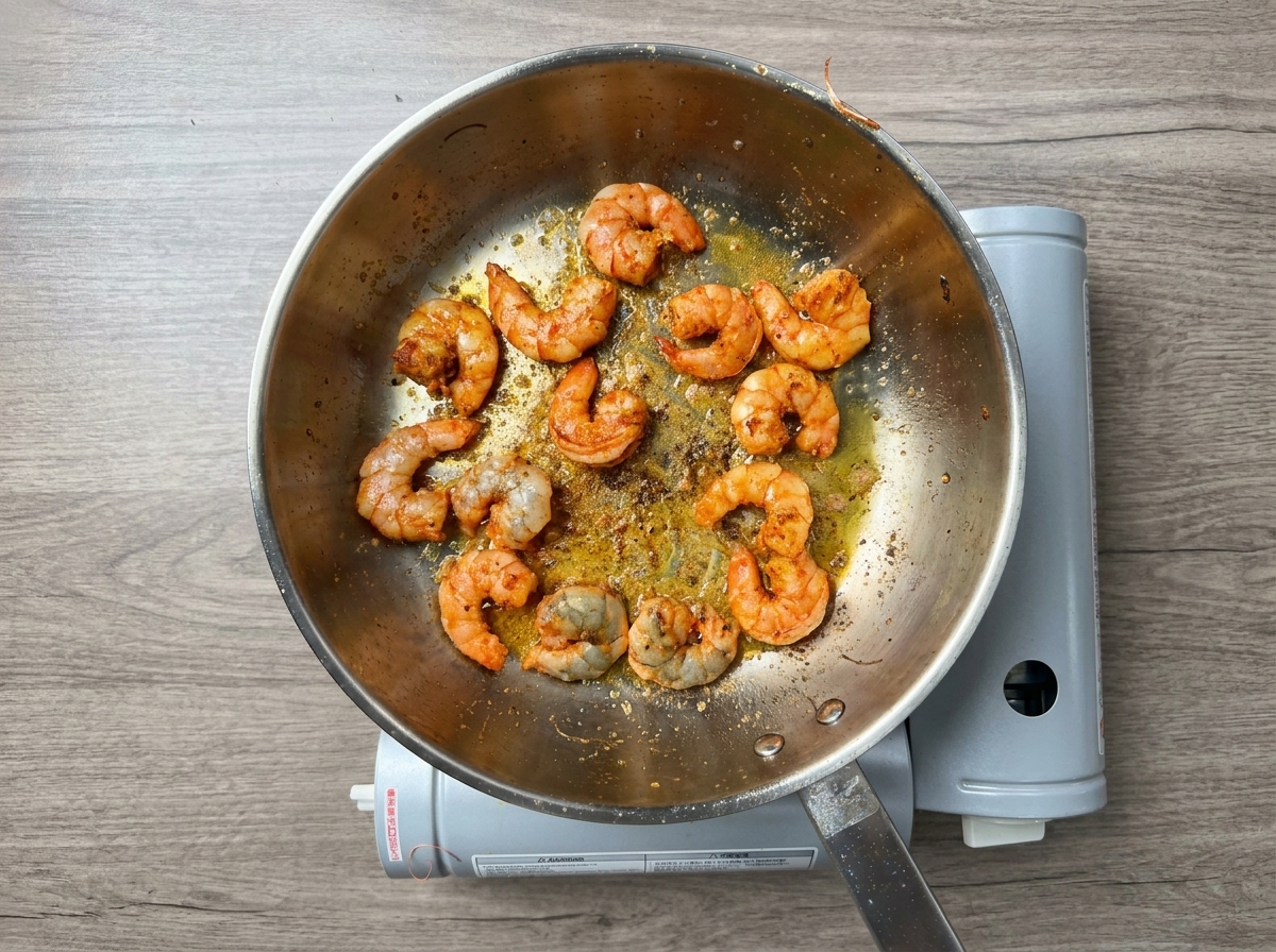 Seasoned peeled shrimp cooking in reddish infused oil in a stainless steel pan.