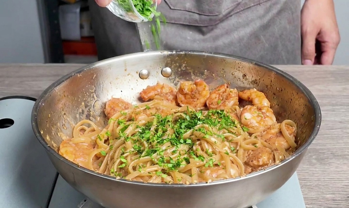 Chopped fresh parsley being sprinkled from a small glass bowl onto linguine and cooked shrimp in a pan.