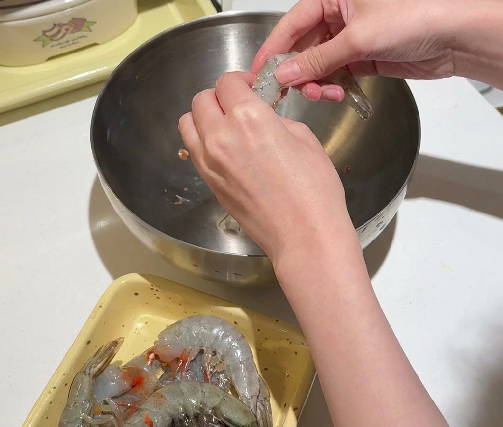 Hands twisting the head and shell off a raw shrimp over a stainless steel bowl.