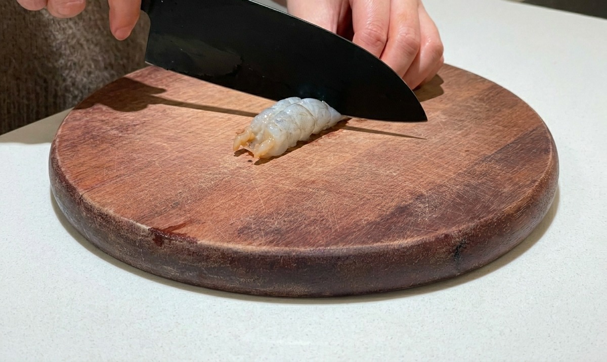 A knife slicing along the back of a peeled raw shrimp on a round wooden cutting board.