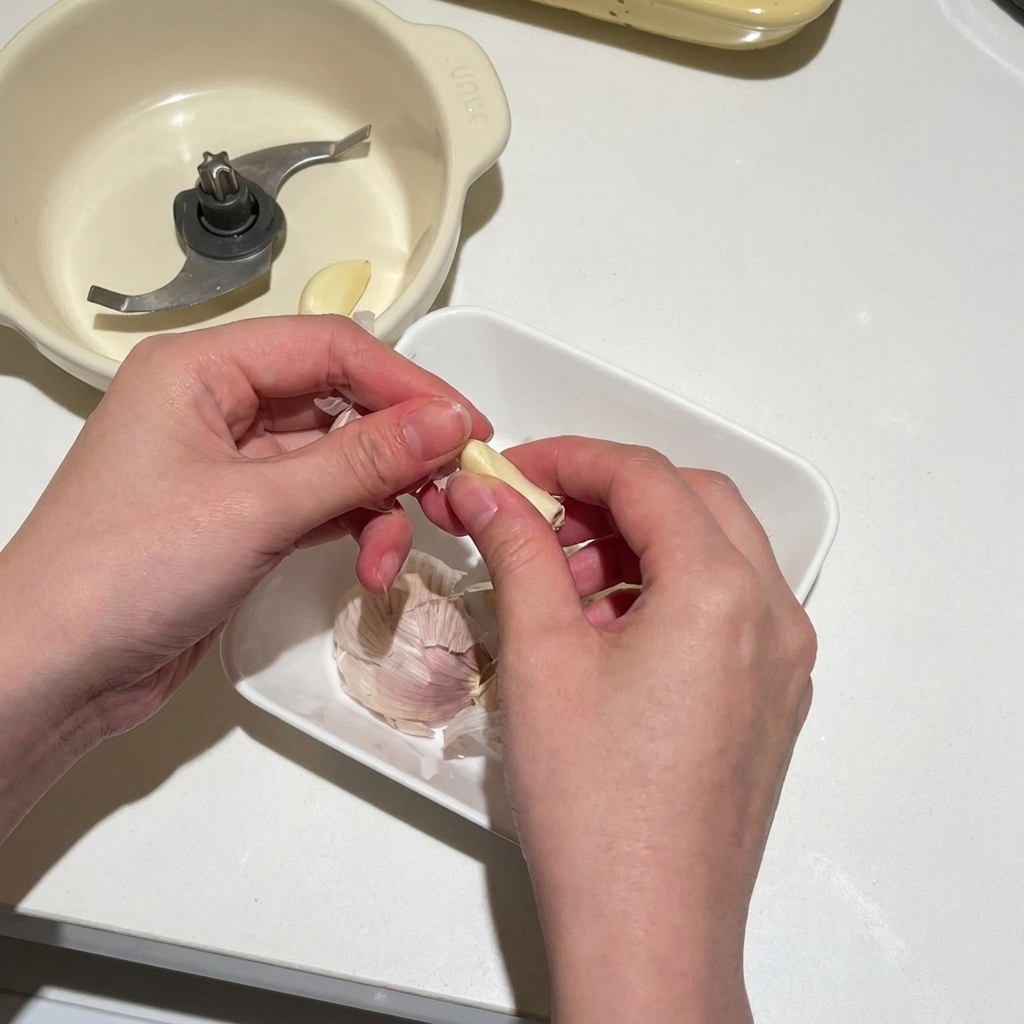 Hands peeling fresh garlic cloves and dropping them into a white container next to an electric food chopper blade.