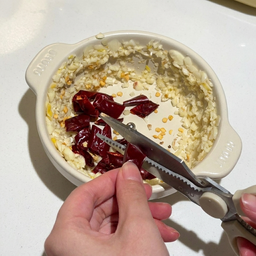 Hands using kitchen scissors to cut dried red chilies into a small bowl of minced garlic.