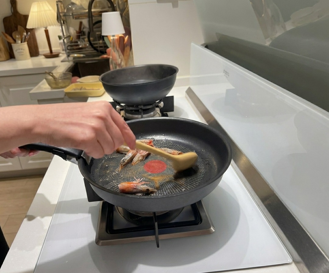 A hand using a wooden spoon to stir-fry raw shrimp heads in hot oil inside a black frying pan.