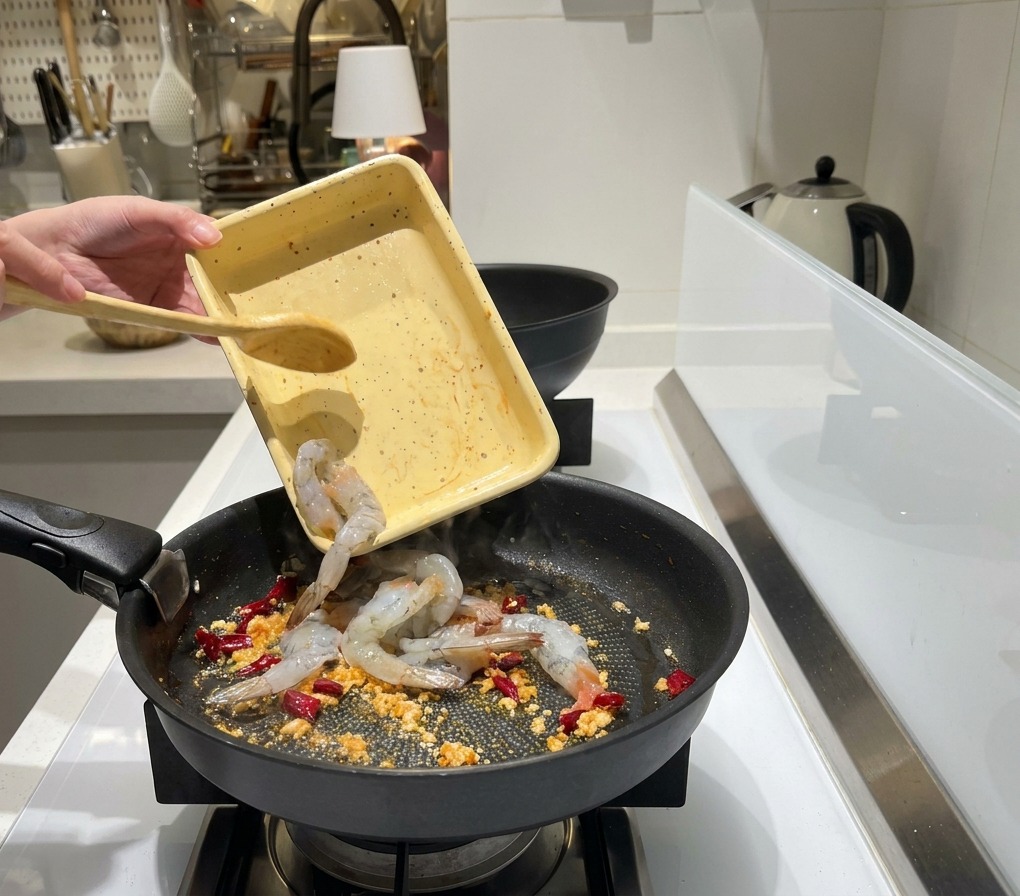 Peeled raw grey shrimp being poured from a small yellow tray into a frying pan with sizzling garlic and chilies.