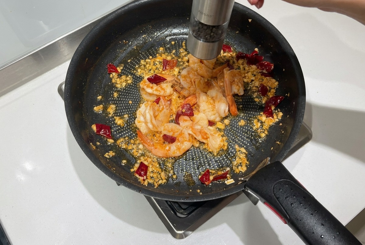 Fresh black pepper being ground from a mill over partially cooked pink shrimp in a frying pan.