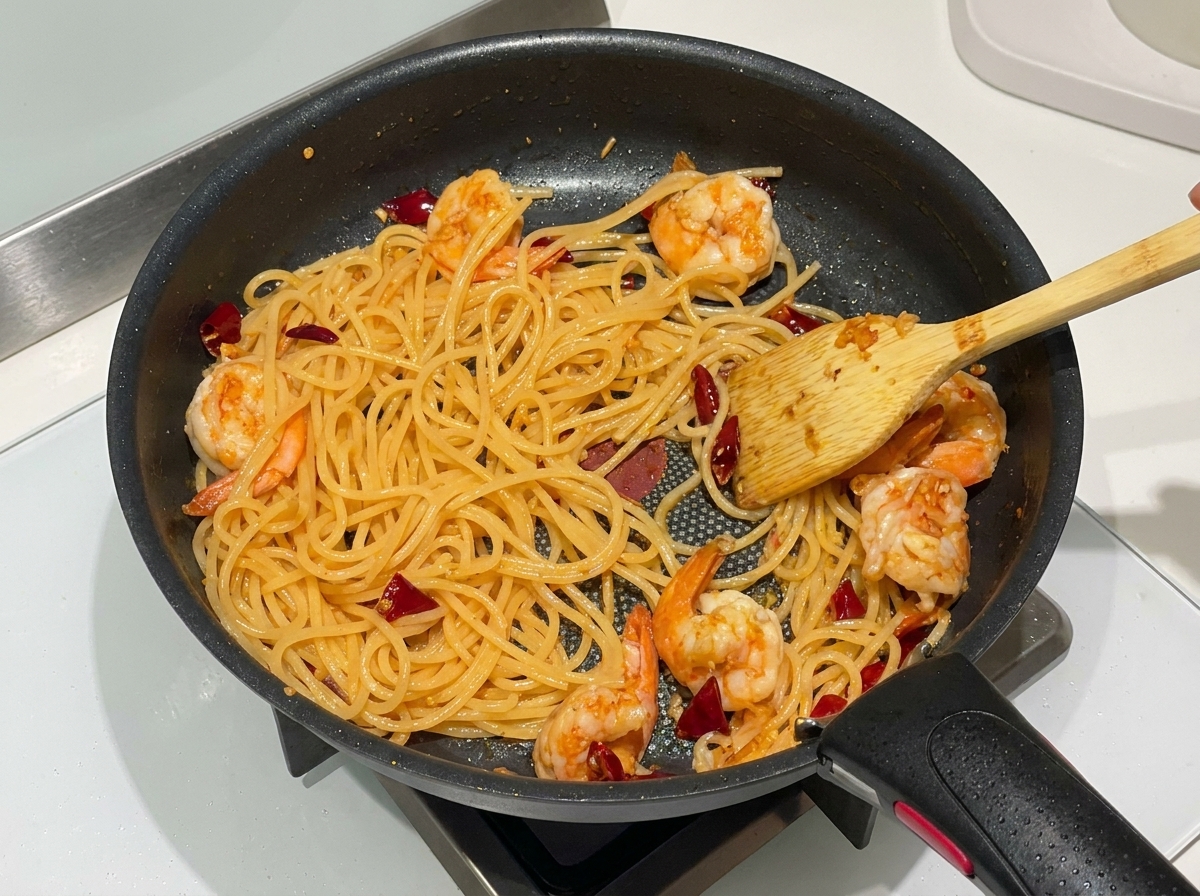 Cooked spaghetti, curled pink shrimp, and dried red chilies being tossed together in a black non-stick skillet with a wooden spatula.