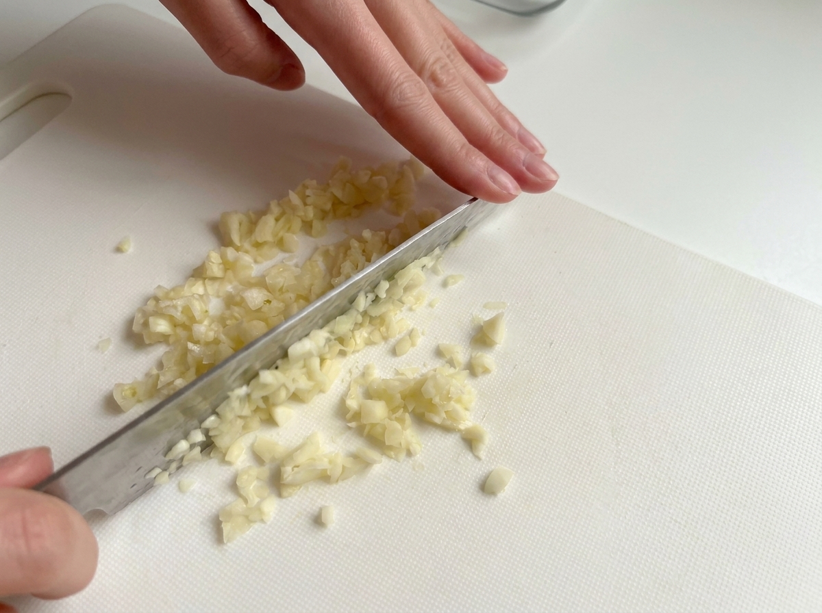 Hands using a knife to finely chop garlic cloves on a white cutting board.