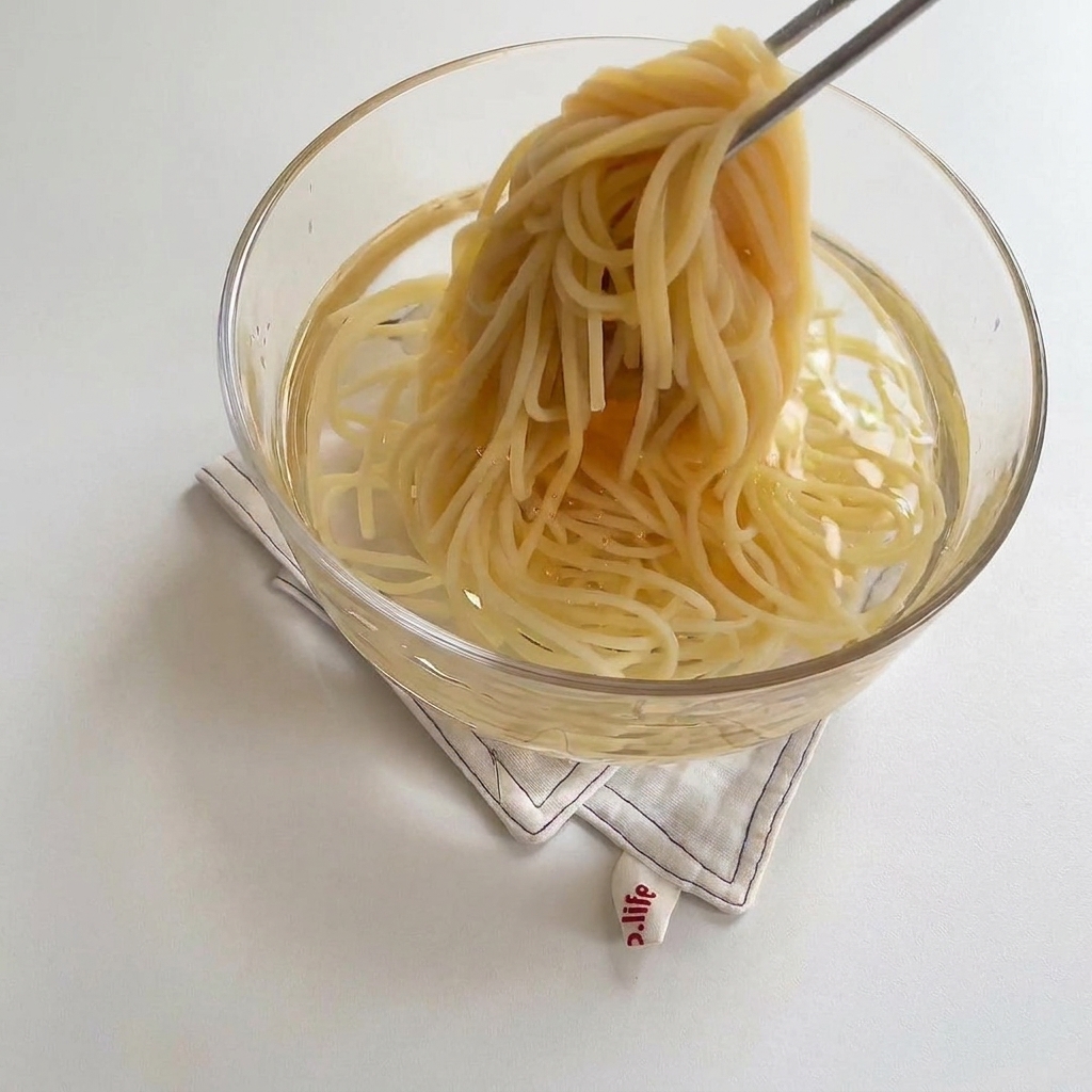 Cooked spaghetti noodles being placed into a clear glass bowl containing cold water.