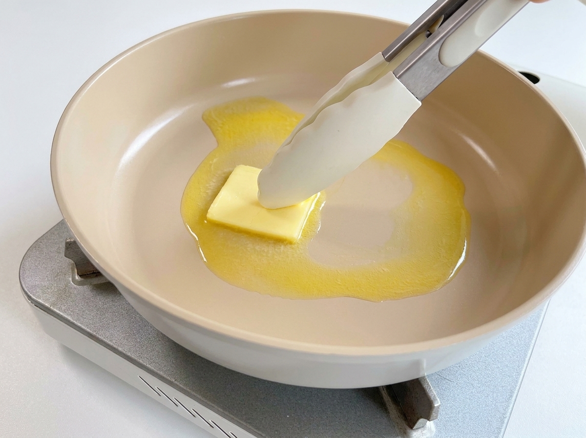 A small square of yellow butter melting in a hot pan, being pushed around by silicone-tipped tongs.
