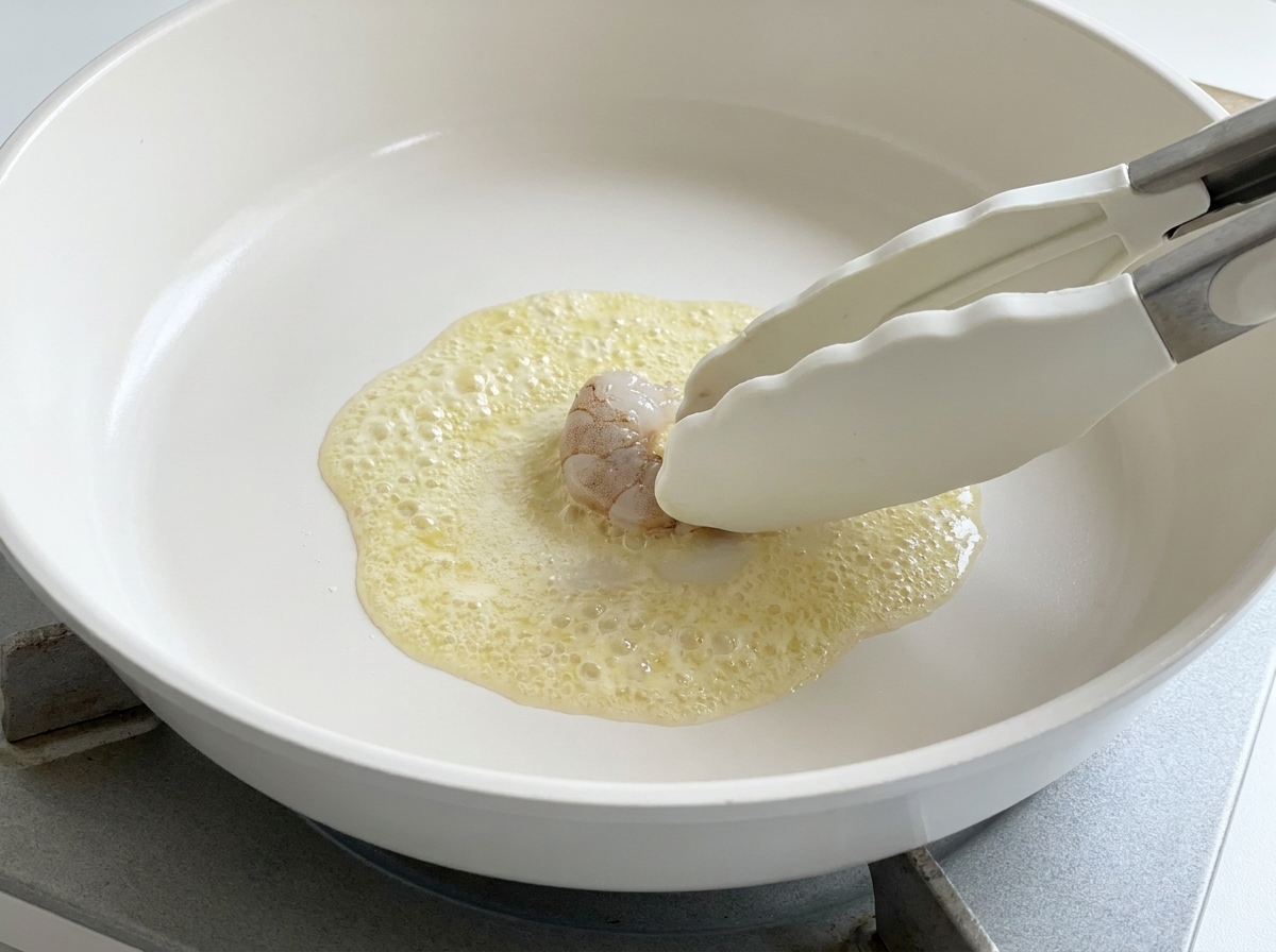A pair of silicone tongs placing a raw prawn into a pan of bubbling melted butter.