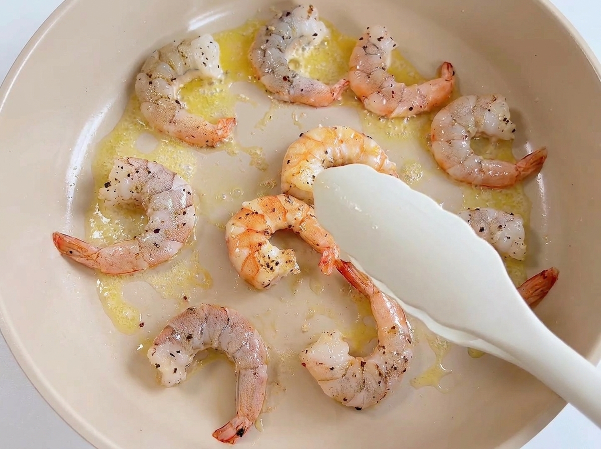 White tongs picking up a cooked pink prawn from a pan filled with other cooked prawns and residual butter.
