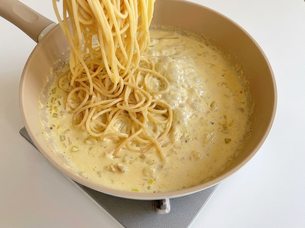 Cooked spaghetti noodles being lowered into a pan of simmering, creamy cheese sauce.