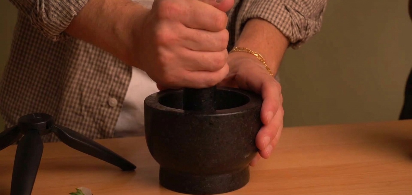 Hands gripping a pestle to firmly crush toasted black peppercorns inside a heavy black stone mortar.