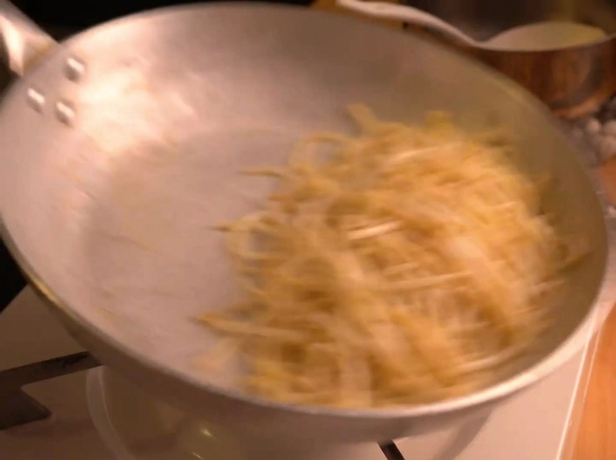 Spaghetti being vigorously tossed in a silver skillet, creating motion blur as the creamy sauce emulsifies.