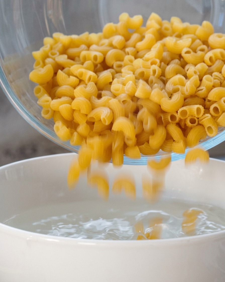 Dry spiral macaroni being poured from a glass bowl into a white pot of hot water.