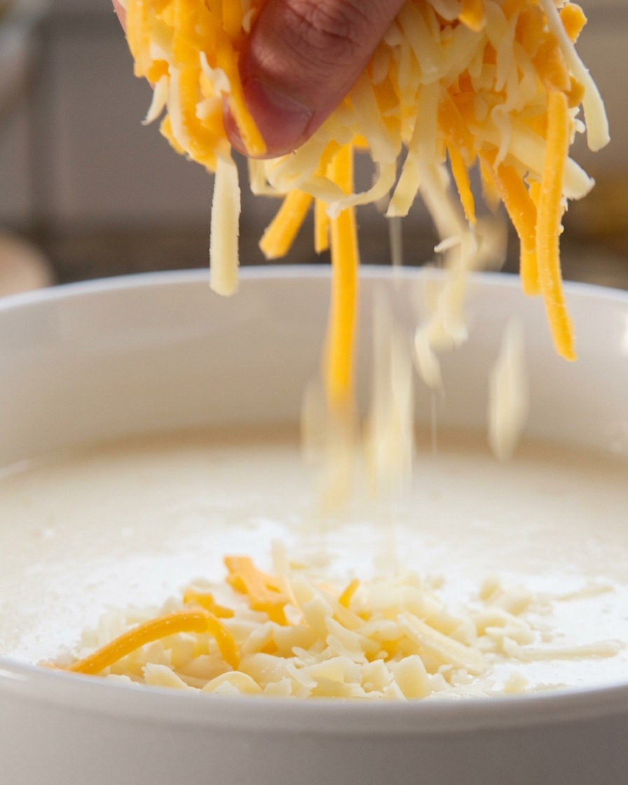 A hand sprinkling a mixture of shredded white and orange cheese into a simmering white liquid.