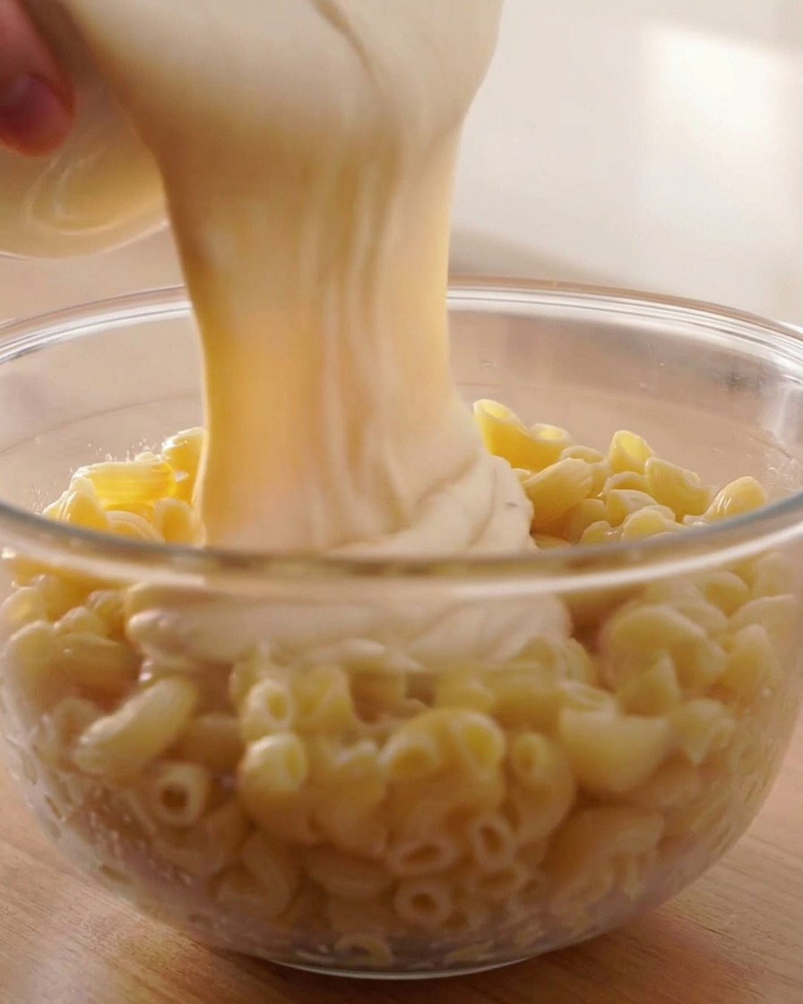 Thick, creamy cheese sauce being poured from a white container over cooked spiral macaroni in a glass bowl.
