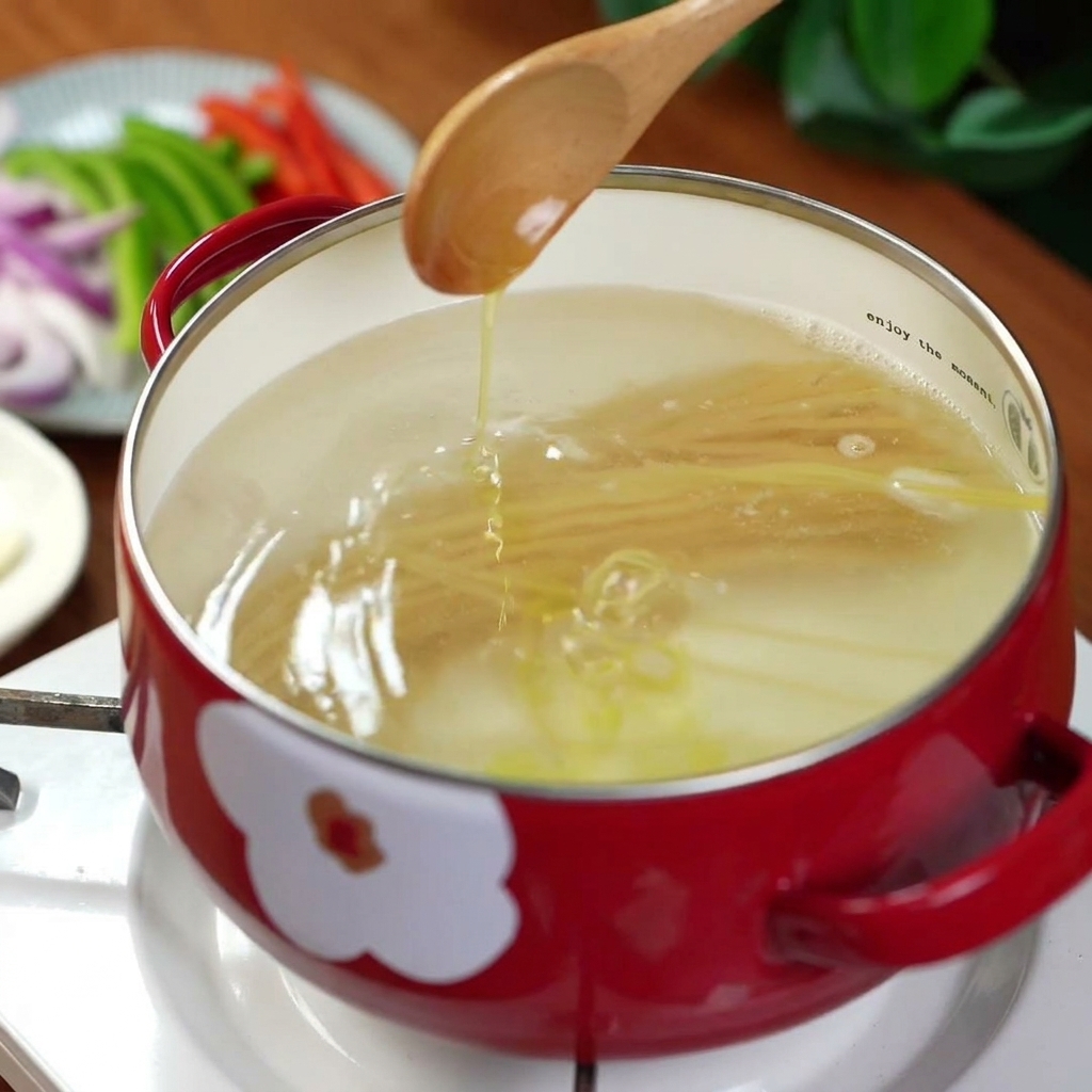 Olive oil being poured from a wooden spoon into a red pot filled with boiling water and spaghetti noodles.