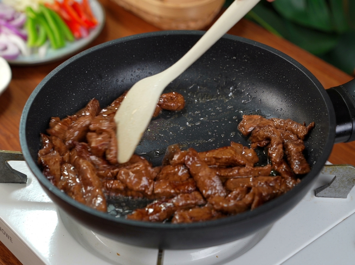 Slices of marinated beef being stir-fried in a hot black non-stick pan with a light-colored silicone spatula.