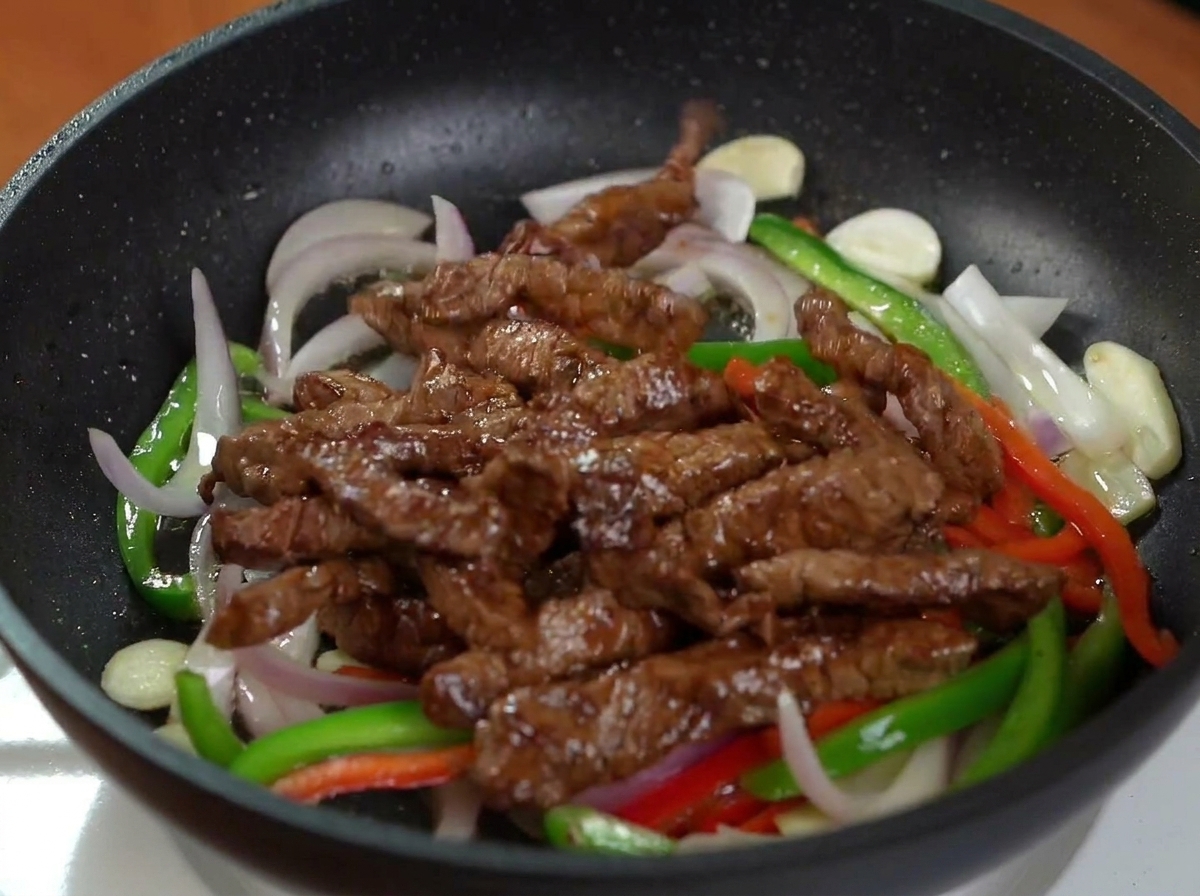 Browned beef strips added to a black frying pan containing sautéed red onions, green bell peppers, and red bell peppers.