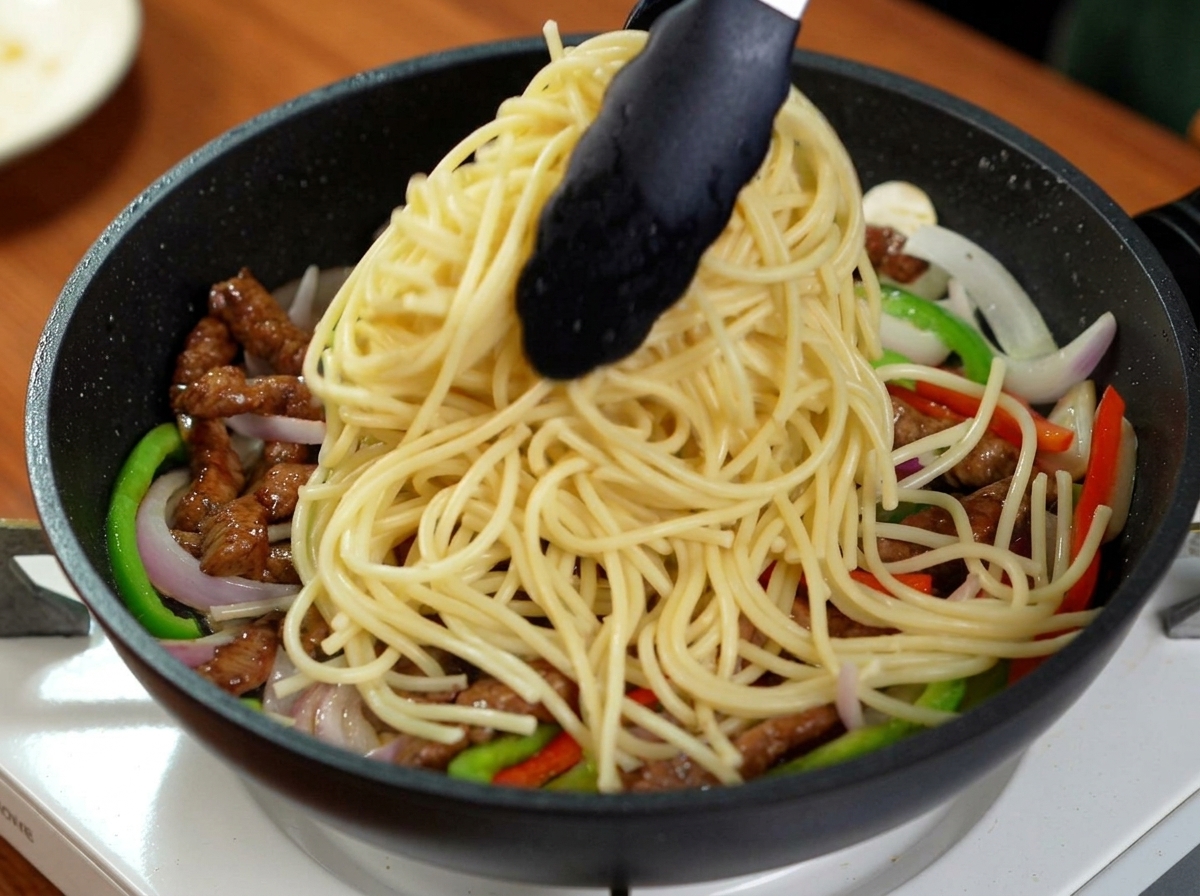Black tongs dropping a large portion of boiled spaghetti into a pan filled with stir-fried beef strips, green bell peppers, and onions.