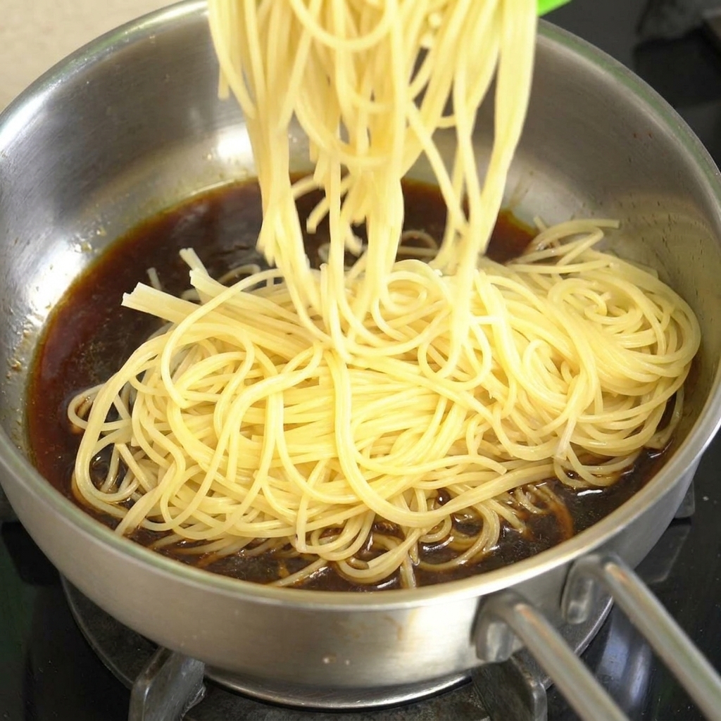 Cooked spaghetti being added to a stainless steel pan filled with dark brown black pepper beef sauce.